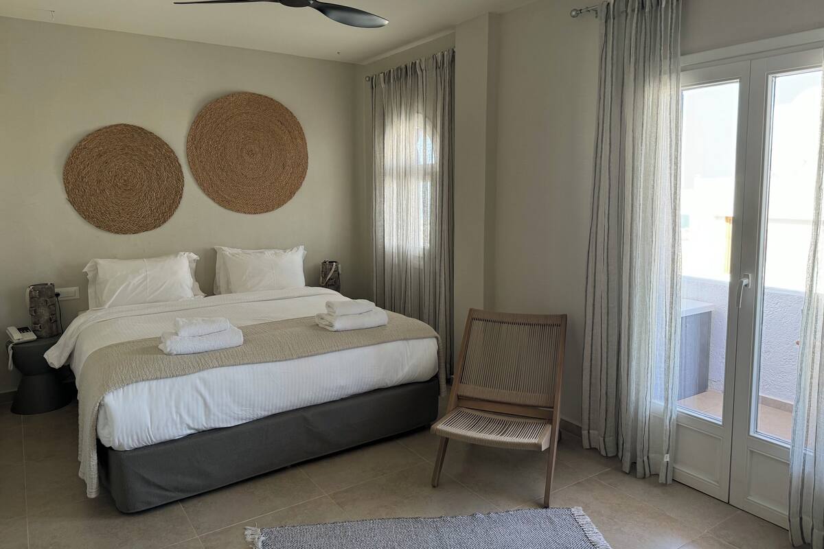Quarto com cama, duas decorações de parede em tecido, ventilador de teto, cadeira e vista através de uma porta de vidro.