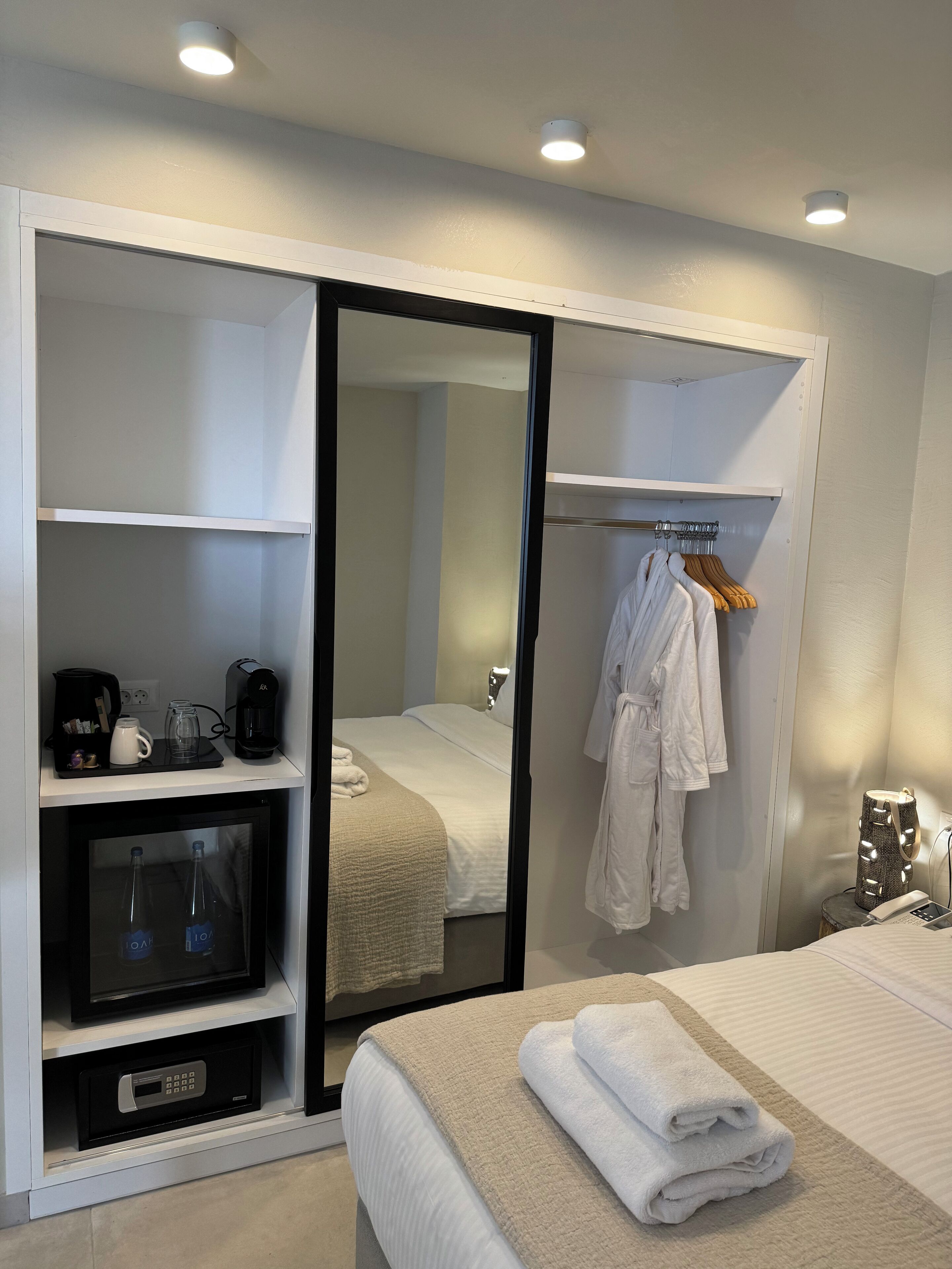 A hotel room with a bed, a mirror, a wardrobe, and a bedside table with a lamp and a phone.