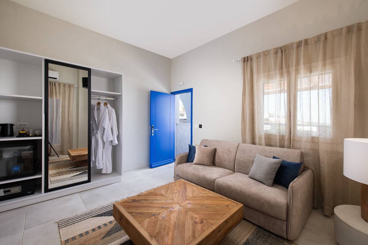 Uma sala de estar moderna com um sofá bege, uma mesa de centro de madeira e um espelho grande. Há uma porta azul e uma janela com cortinas leves.