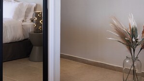 A modern bedroom with a large mirror, a bed with white linens, a bedside table with a lamp, and a vase with dried plants.