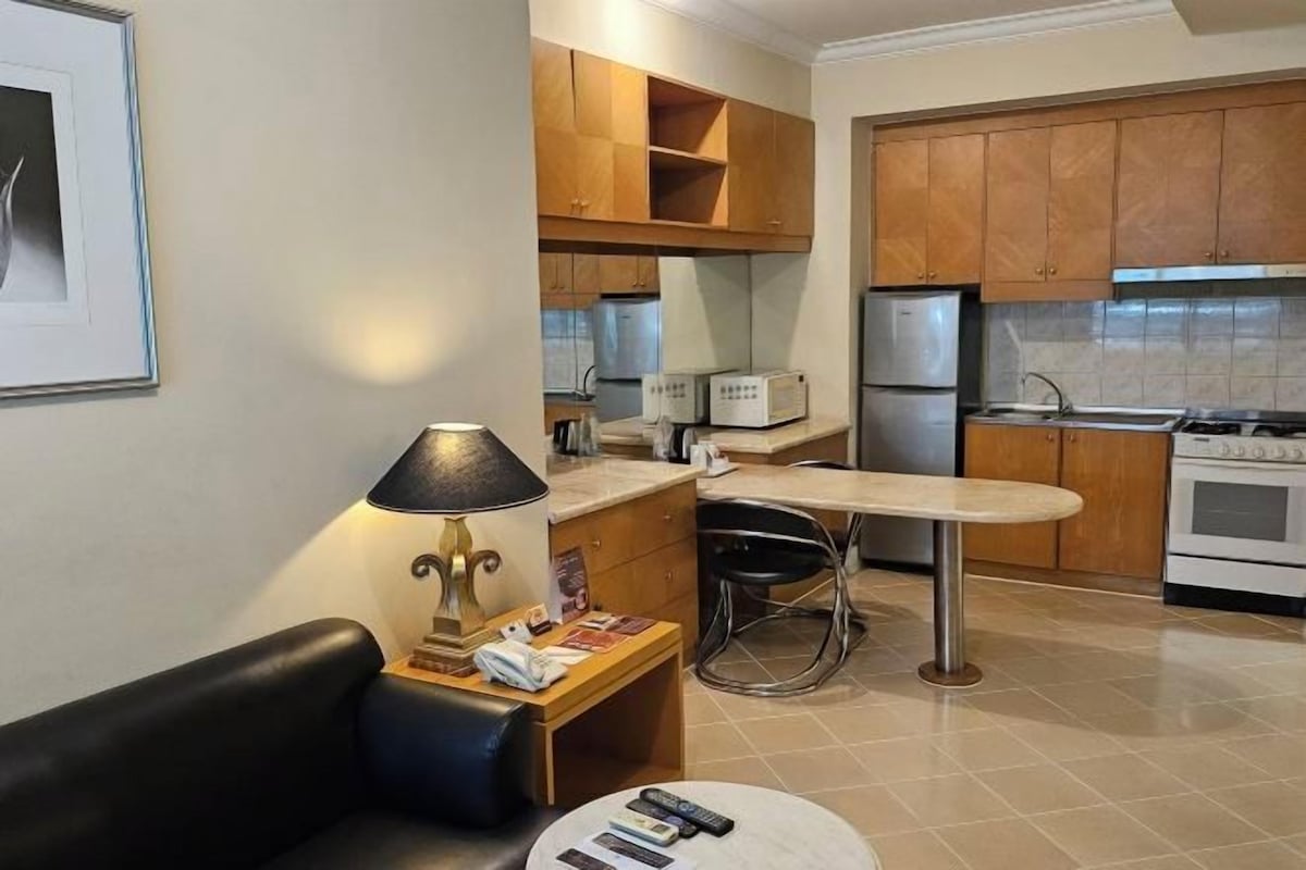 A compact kitchen with wooden cabinets, a dining table, a bar stool, a sofa, and a lamp.