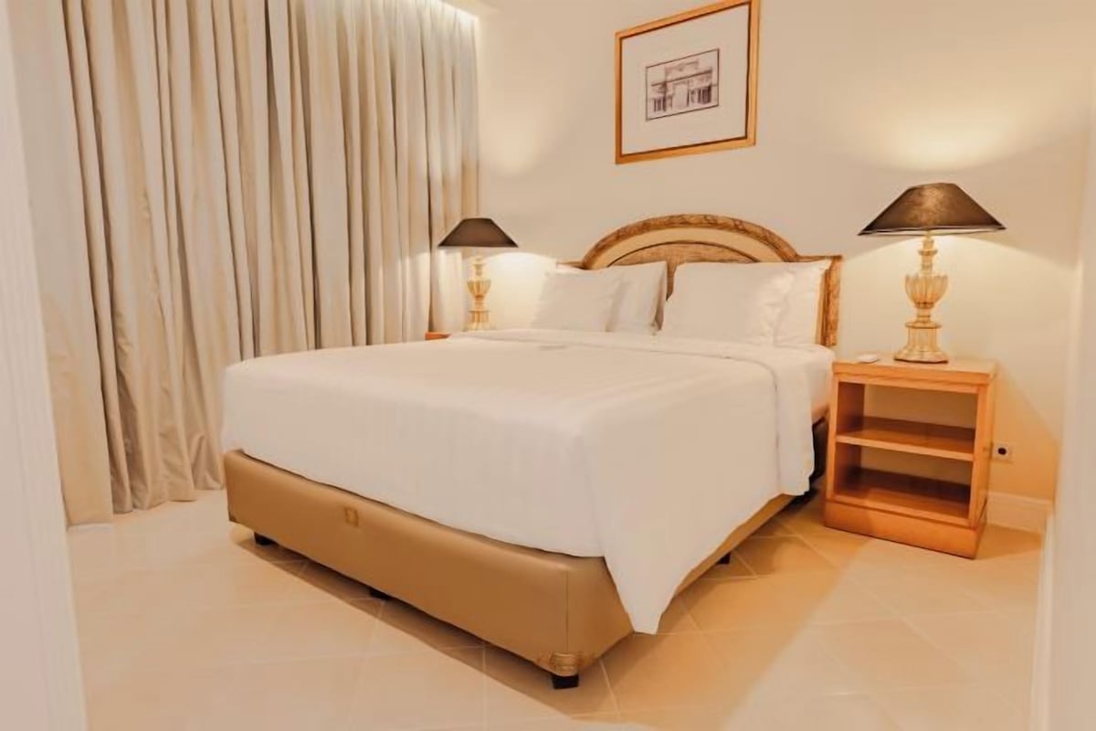 A hotel room with a bed, two bedside tables, a nightstand, a lamp, a curtain, and a framed picture on the wall.