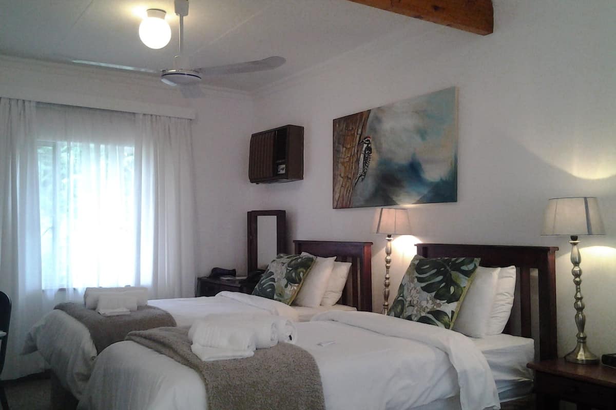 Ein Hotelzimmer mit zwei Betten, einem Deckenventilator, einem Gemälde, einer Nachttischlampe und einem fenster mit Vorhängen.