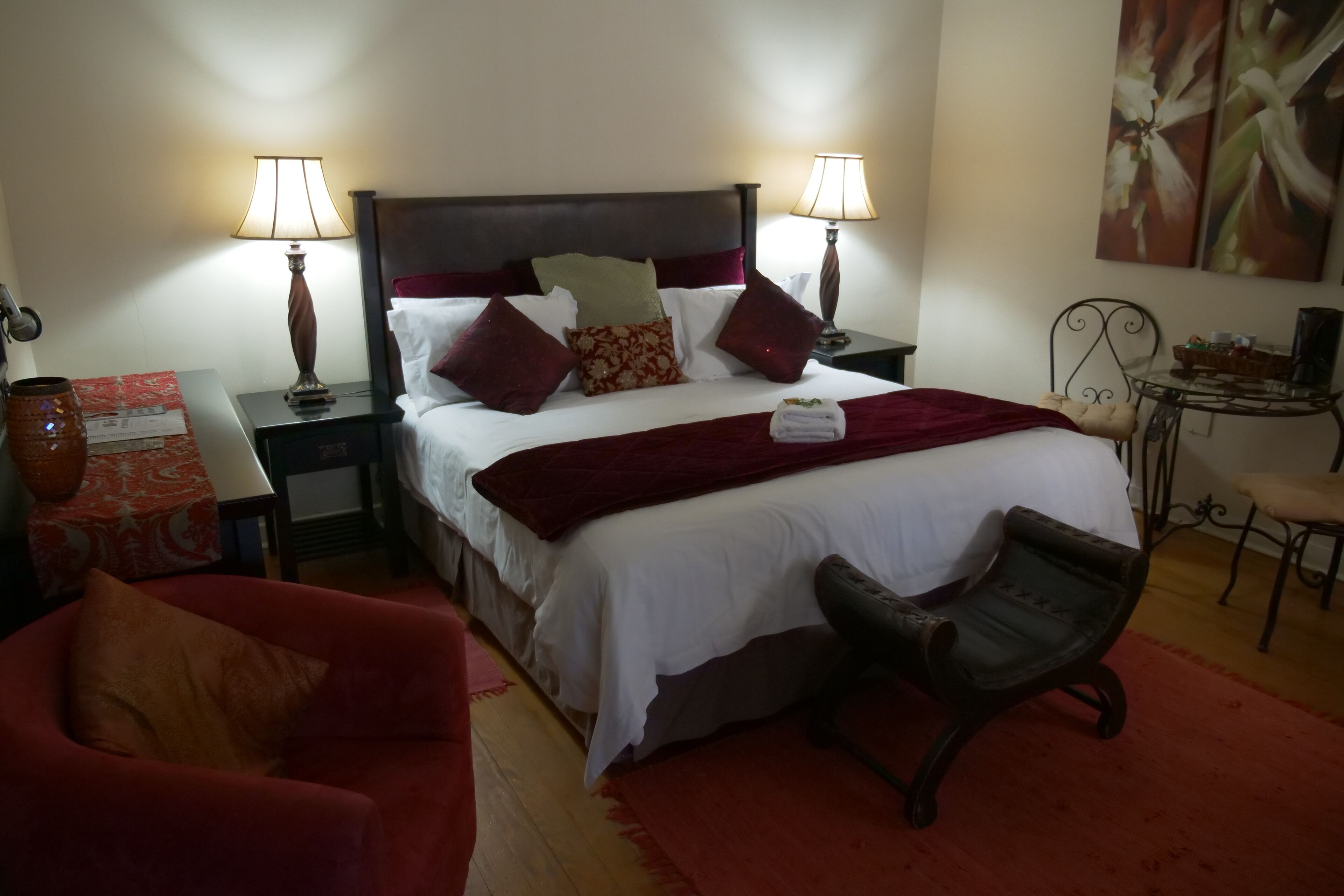 A hotel room with a bed, two bedside lamps, a red armchair, a small table with a vase, and a painting on the wall.