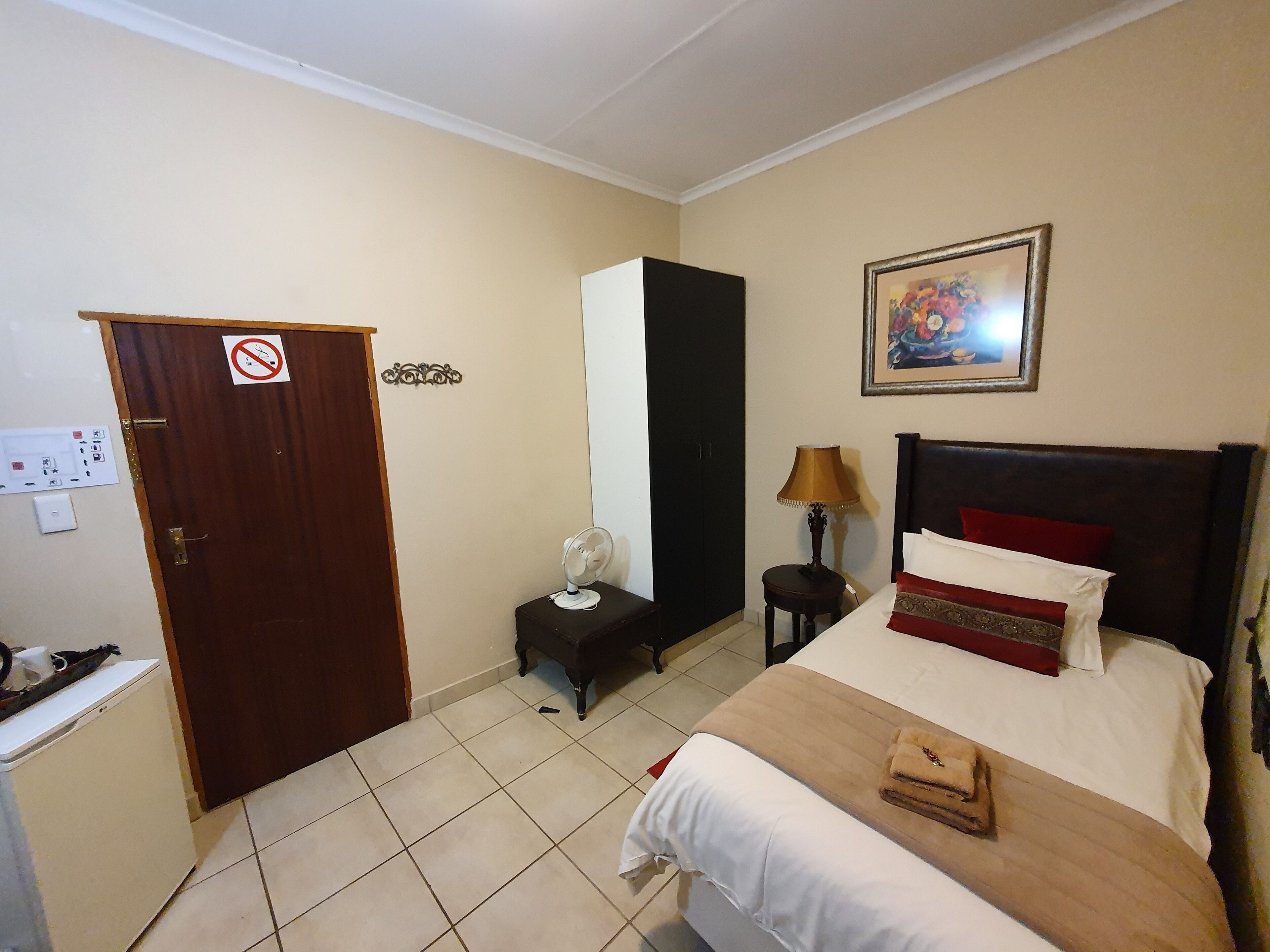 A hotel room with a bed, bedside table, lamp, fan, and a door with a no-smoking sign.