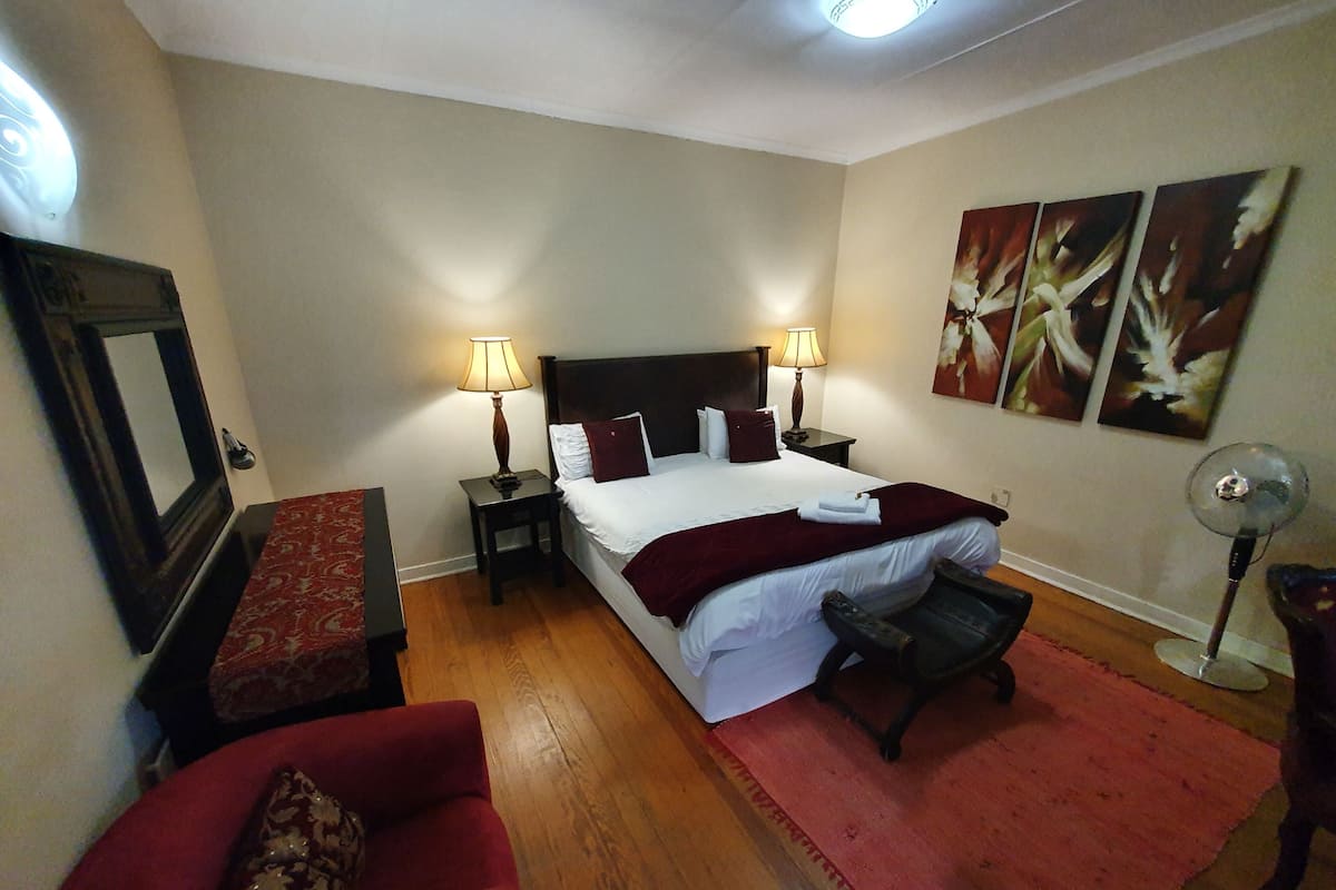 Habitación de hotel con una cama, dos lámparas de noche, una silla, una mesita, un espejo y cuadros en la pared.