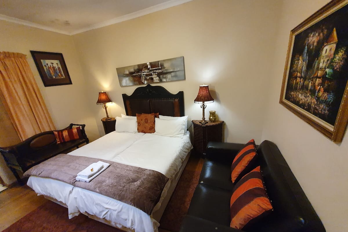 Un cuarto de hotel con una cama, un sofá de cuero negro, dos mesitas de noche con lámparas, un cuadro en la pared y una fotografía enmarcada sobre la cama.