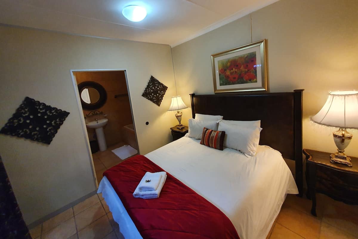 Habitación de hotel con cama, mesita de noche, lámpara, un cuadro, un espejo y un baño visible a través de una puerta abierta.