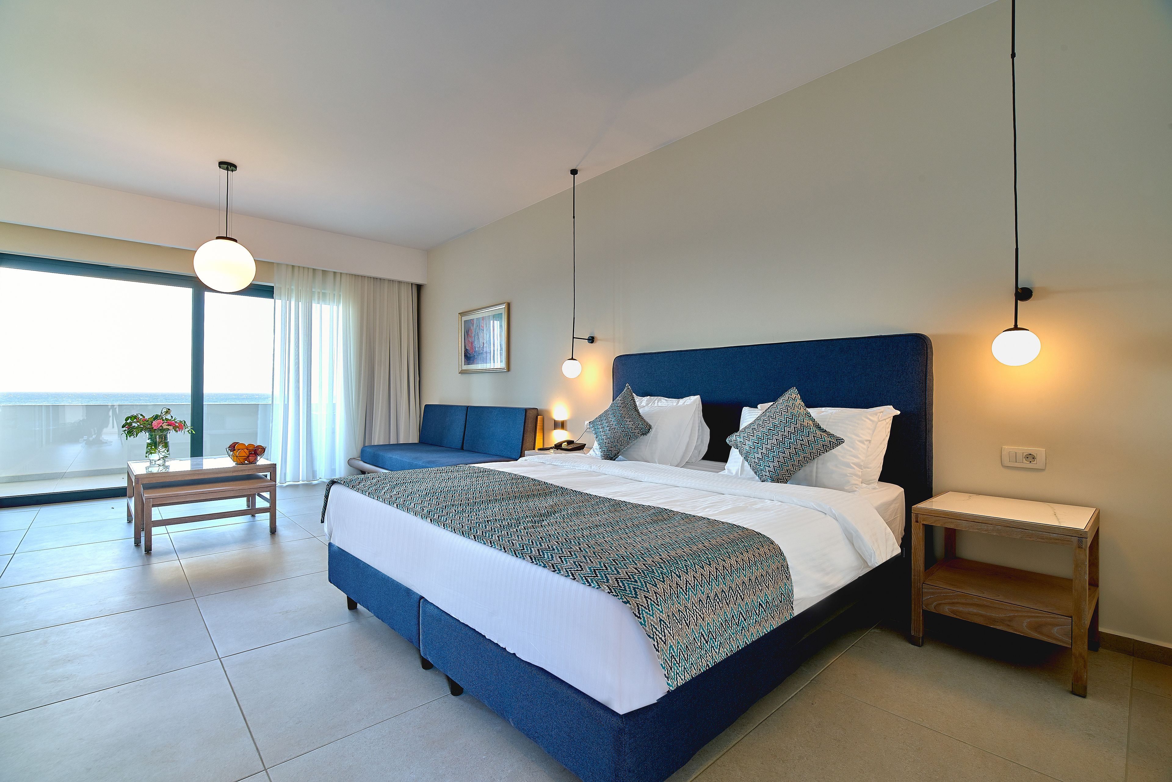 A modern bedroom with a large bed, bedside tables, a small table with flowers, and a view of the outdoors.