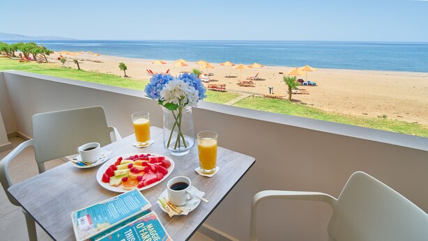 Balcón con mesa lista para el desayuno, con vista a una playa con sombrillas y sillas.