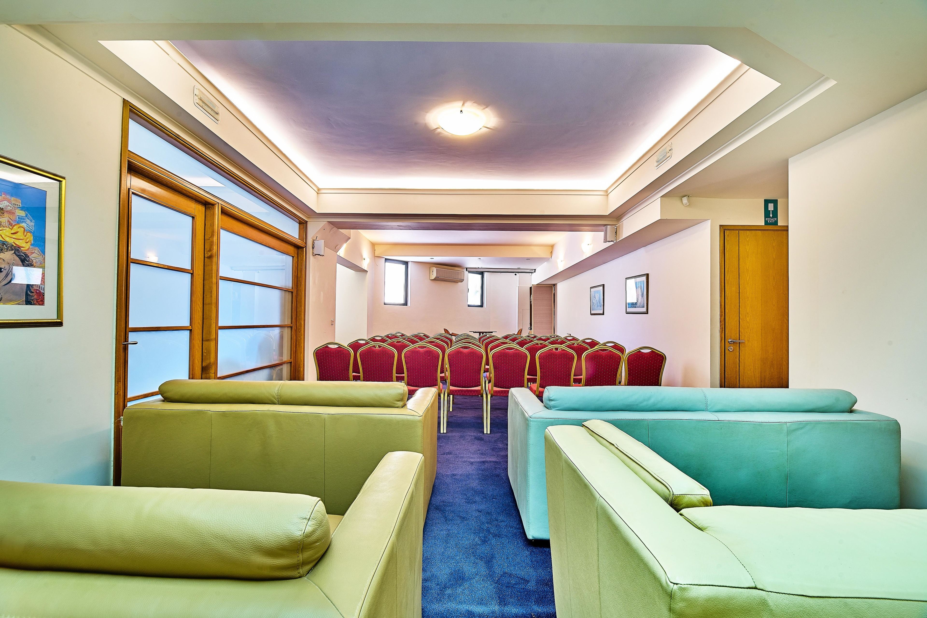 Habitación de hotel con un largo pasillo, filas de sillas rojas y dos filas de sofás color beige y verde azulado.