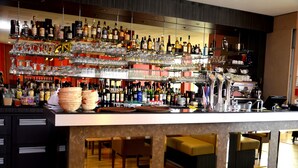 Uma área de bar com várias prateleiras de garrafas e copos, um balcão com banquetas e uma área de estar com mesas e cadeiras.