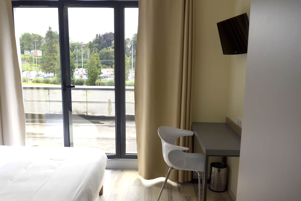 A hotel room with a bed, a desk, a chair, a TV, and a view of a parking lot and trees.
