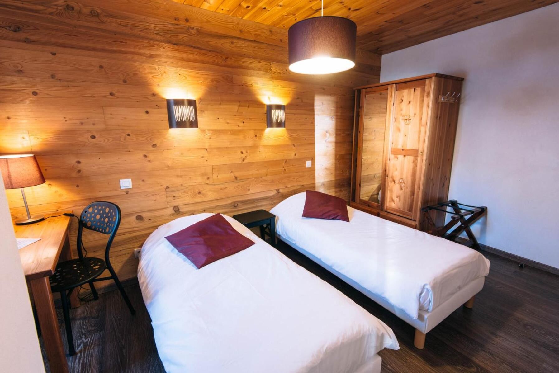 A room with two single beds, a wooden wardrobe, a desk with a chair, and a hanging light fixture.