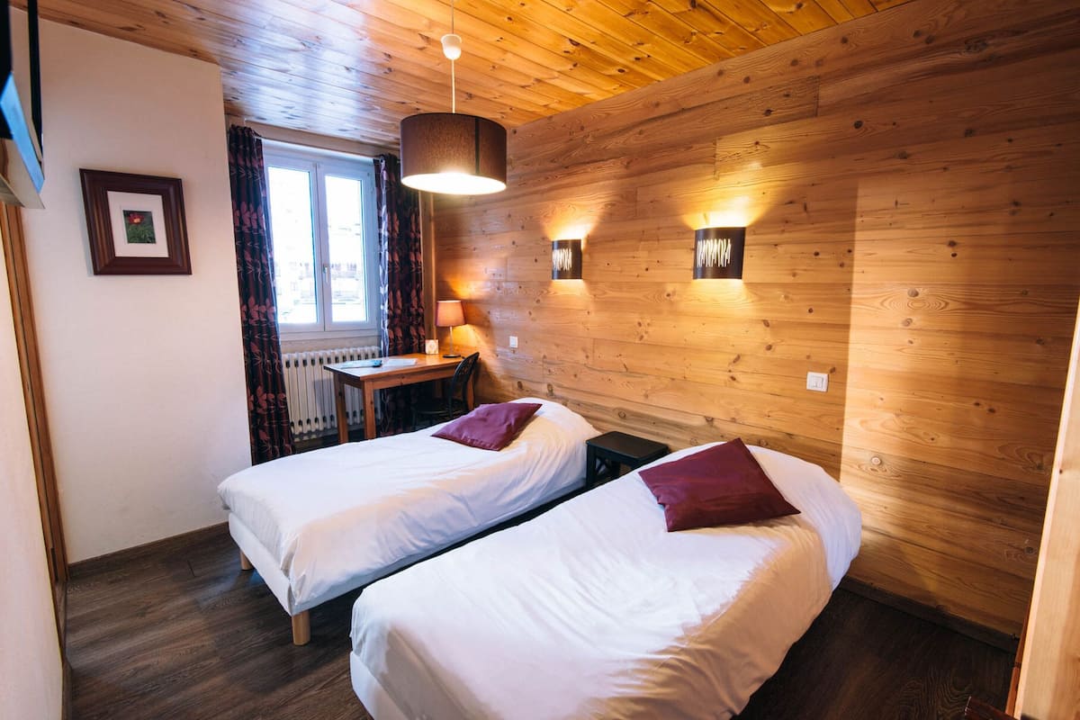 A cozy room with two single beds, a wooden ceiling, a table with a lamp, and a window with curtains.