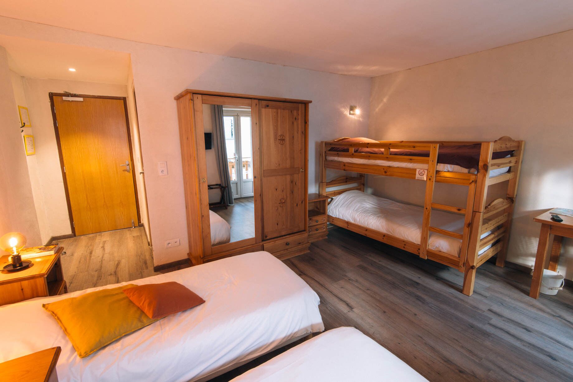 Une chambre avec deux lits simples, un lit superposé, une armoire en bois, un miroir et une petite table avec une lampe.