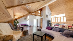A spacious loft with high ceilings, exposed wooden beams, a cozy seating area with a sofa and armchair, a dining table with chairs, and a large window providing natural light. - Hôtel Châlet de Lanza (Abriès-Ristolas)