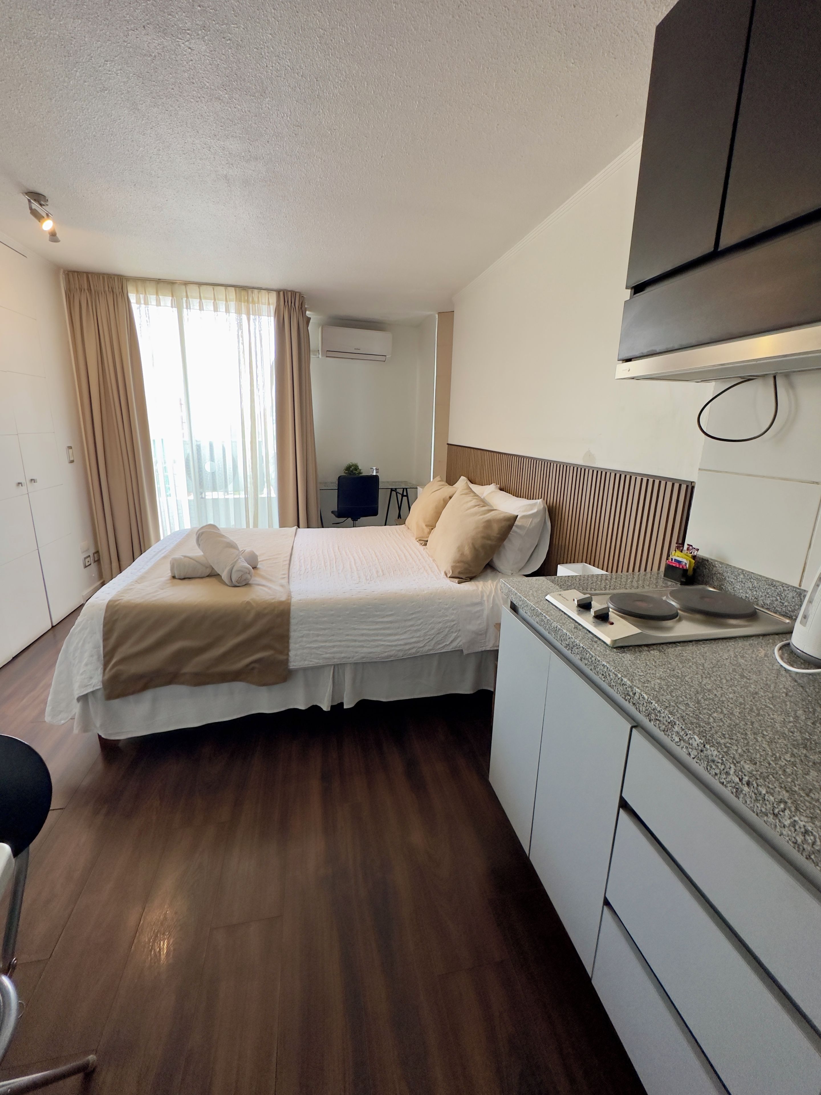 Una habitación de hotel con cama, escritorio, silla, kitchenette y aire acondicionado.