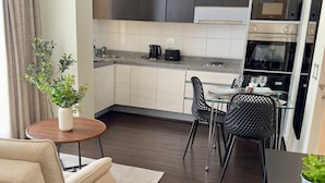 A modern kitchen with dark cabinets, a dining area with a glass table and chairs, and a cozy seating area with a sofa and a small side table. - Rentasuite (Santiago)