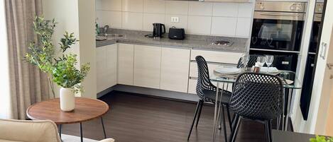 Una cocina moderna con armarios oscuros, un comedor con mesa y sillas de vidrio, y una zona de estar acogedora con un sofá y una pequeña mesita auxiliar.