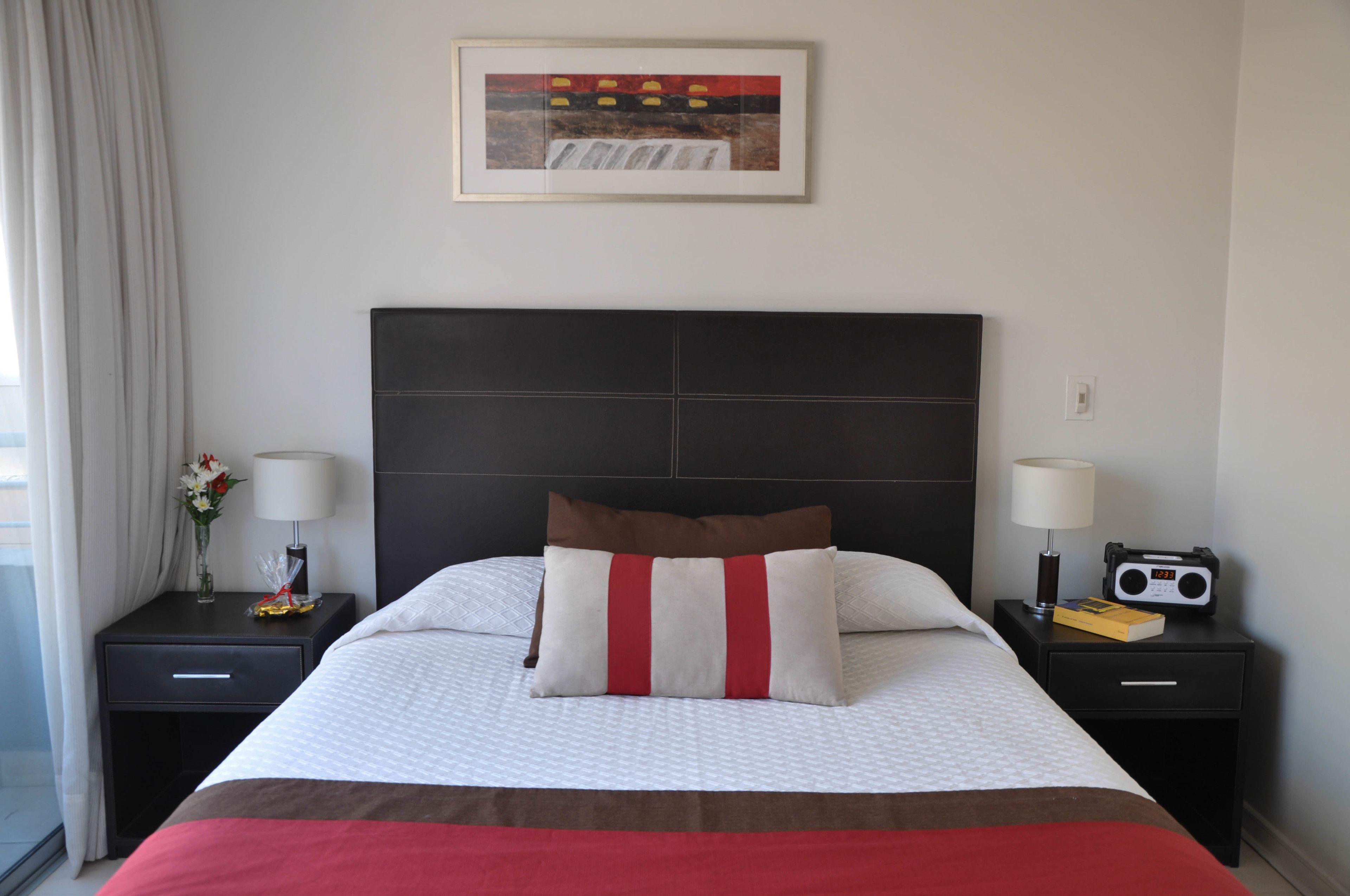 Ein Hotelzimmer mit einem großen Bett, einem gerahmten Bild an der Wand, einem Nachttisch mit Lampe, einem Radio und einer Vase mit Blumen.