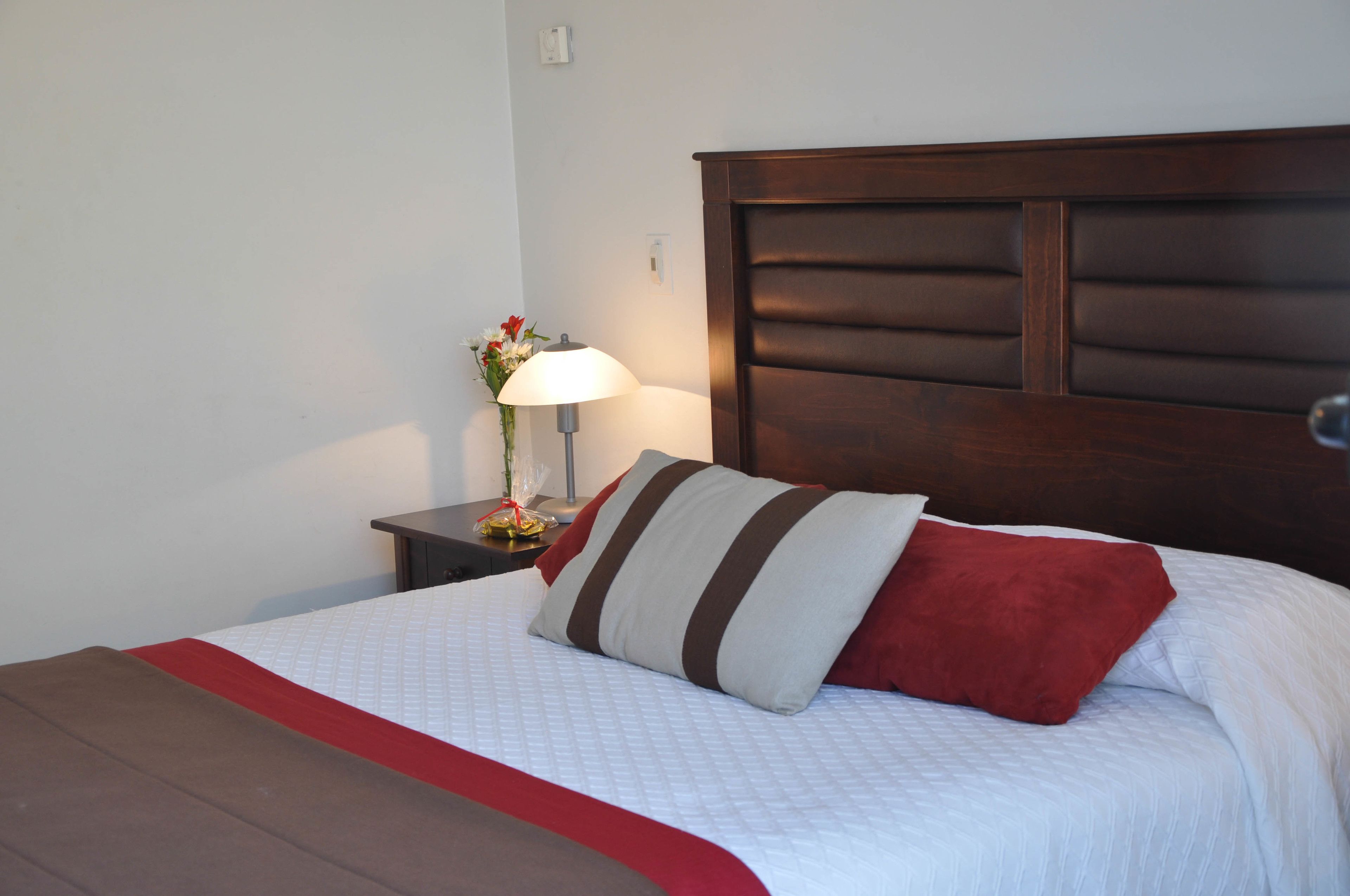 A neatly made bed with a red and white bedspread, a wooden headboard, and a bedside table with a lamp and flowers.