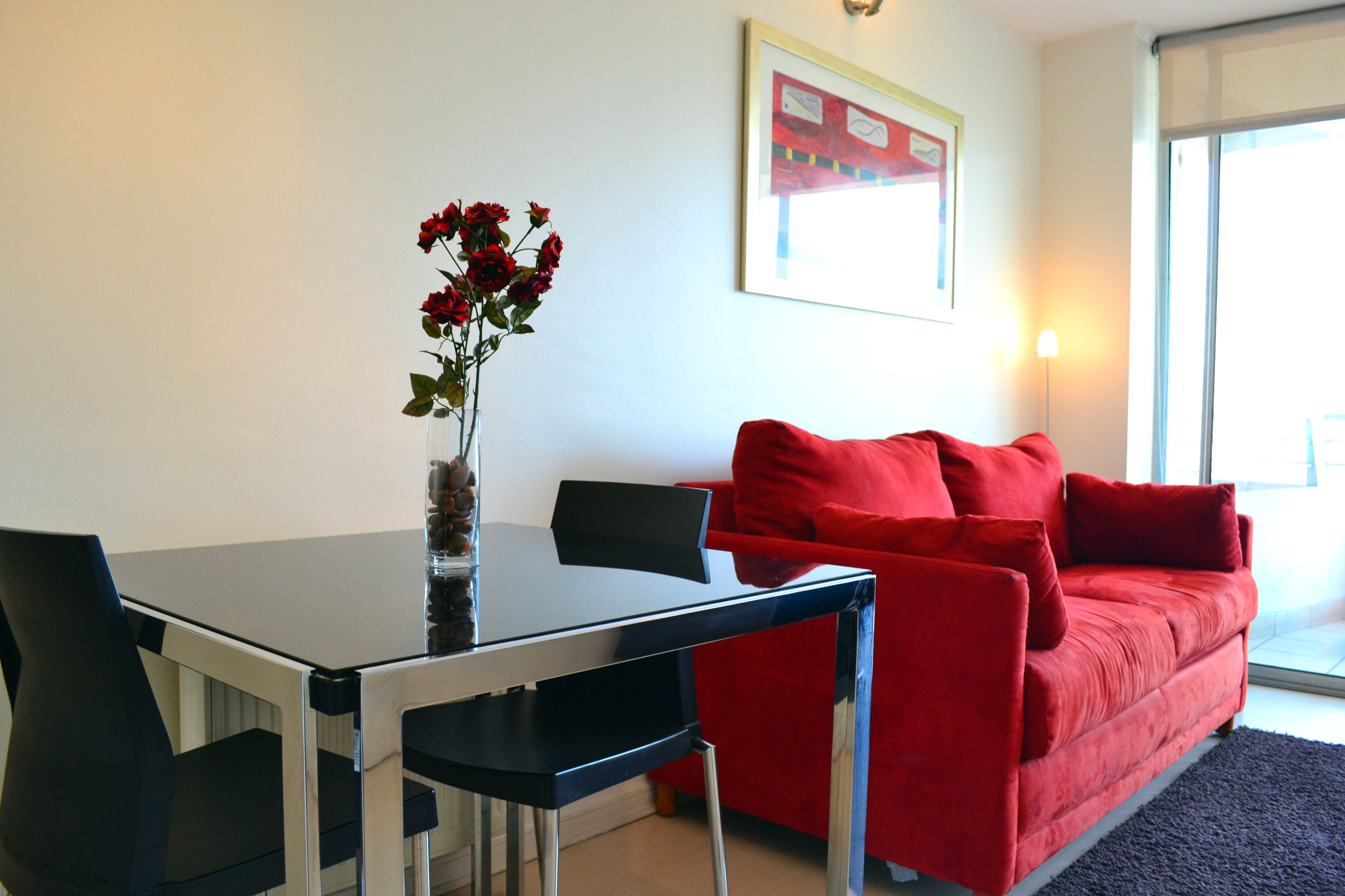 A modern living room with a glass-top dining table, a red sofa, and a vase with red flowers.