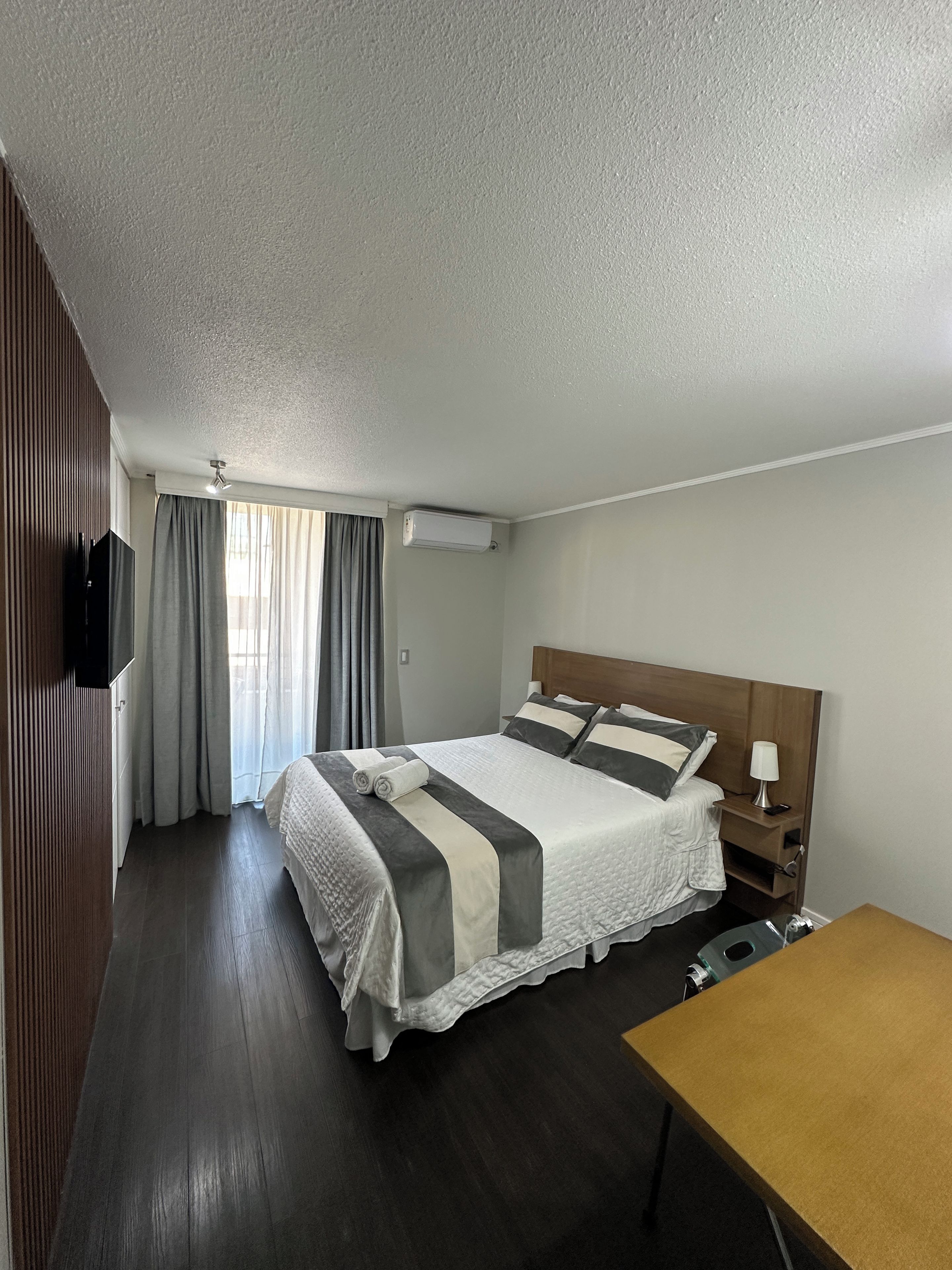 A hotel room with a bed, bedside table, lamp, and a wooden desk.