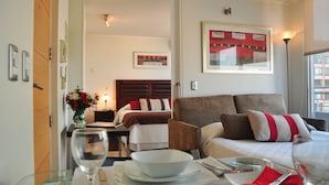 A hotel room with a bed, a sofa, a dining table with a glass top, and a vase with flowers. - Rentasuite (Santiago)