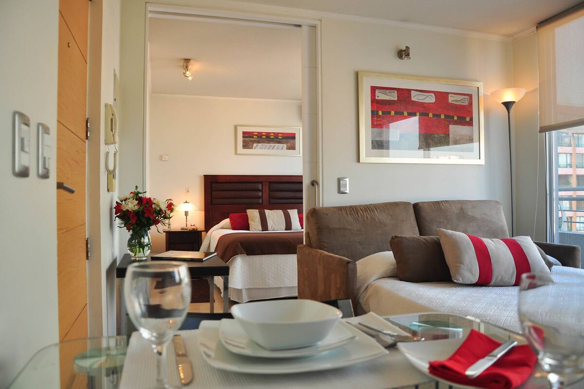 A hotel room with a bed, a sofa, a dining table with a glass top, and a vase with flowers.