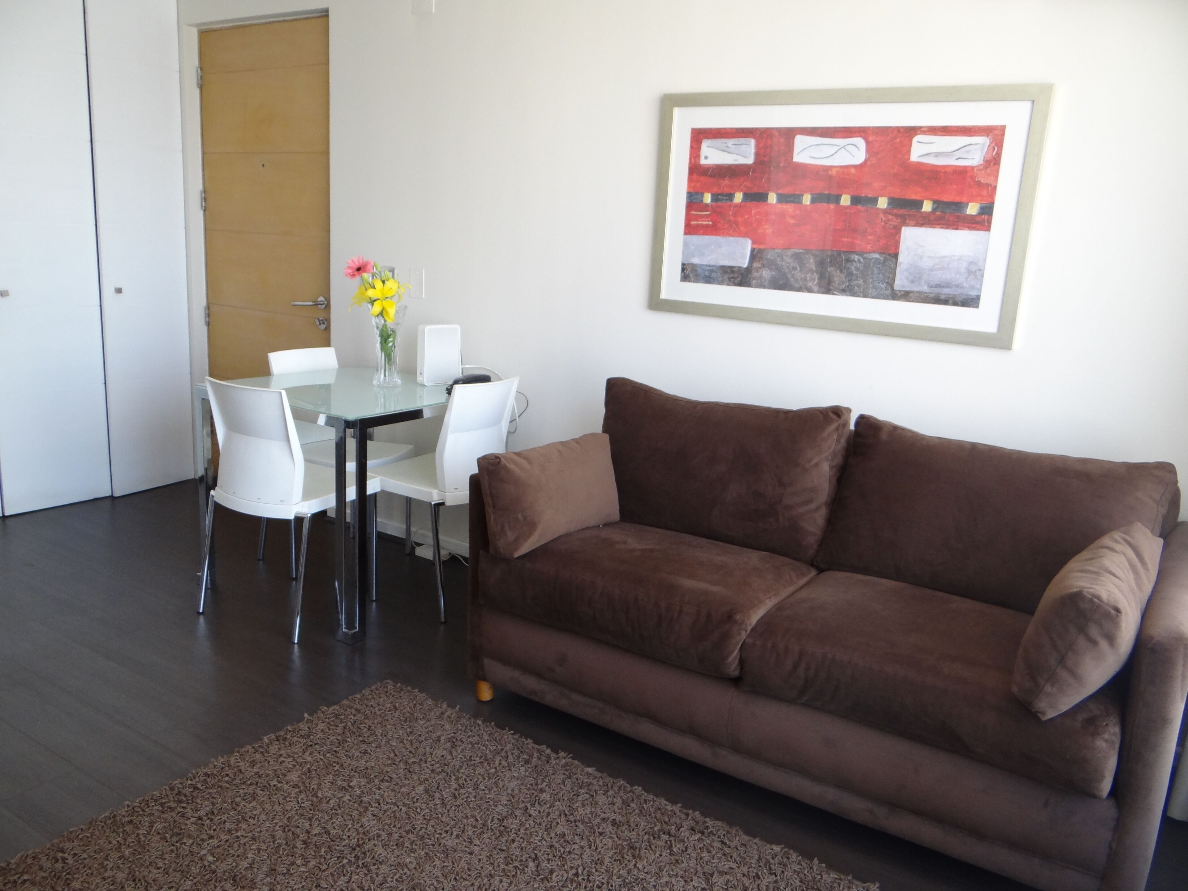 A living room with a brown sofa, a glass table with chairs, a vase with flowers, and a framed picture on the wall.