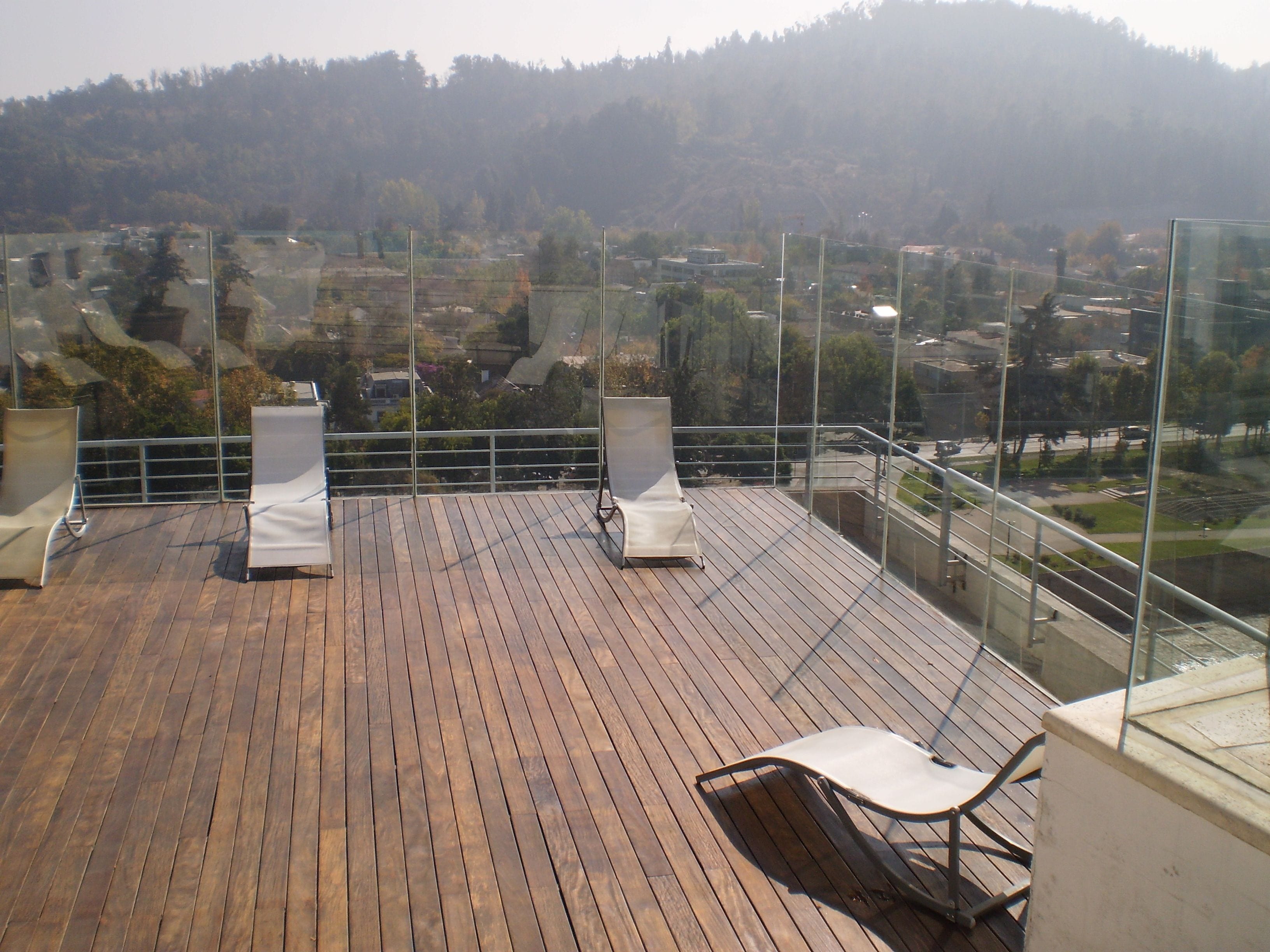 A wooden deck with two white lounge chairs and a glass balustrade overlooking a hilly landscape.