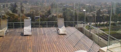 Une terrasse en bois avec deux chaises longues blanches et une balustrade en verre donnant sur un paysage vallonné.
