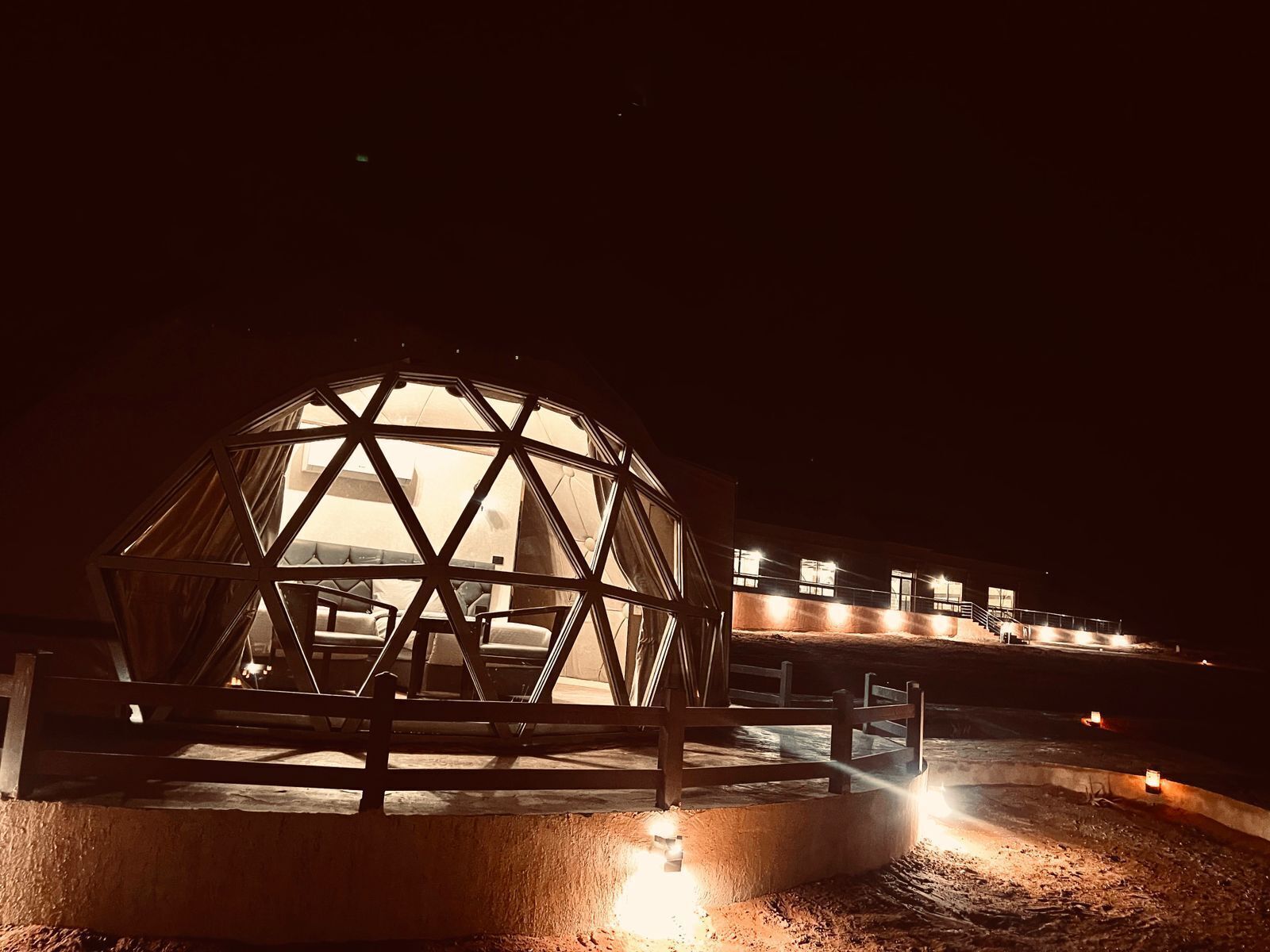 Una estructura de domo geodésico con base de hormigón, rodeada por una valla de madera e iluminada por luces en el suelo.