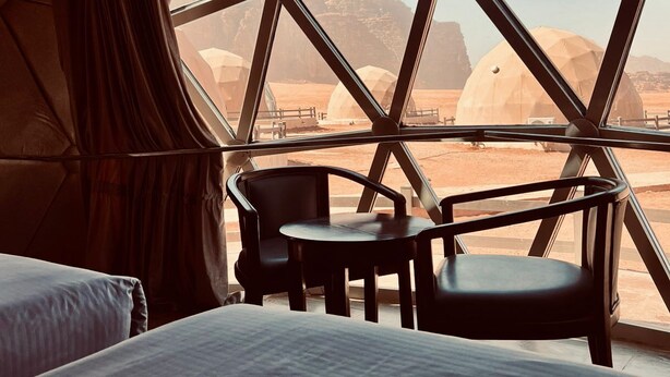 A room with a bed, two chairs, and a large window with a view of a desert landscape. - Beyond Wadi Rum Camp (Wadi Rum)