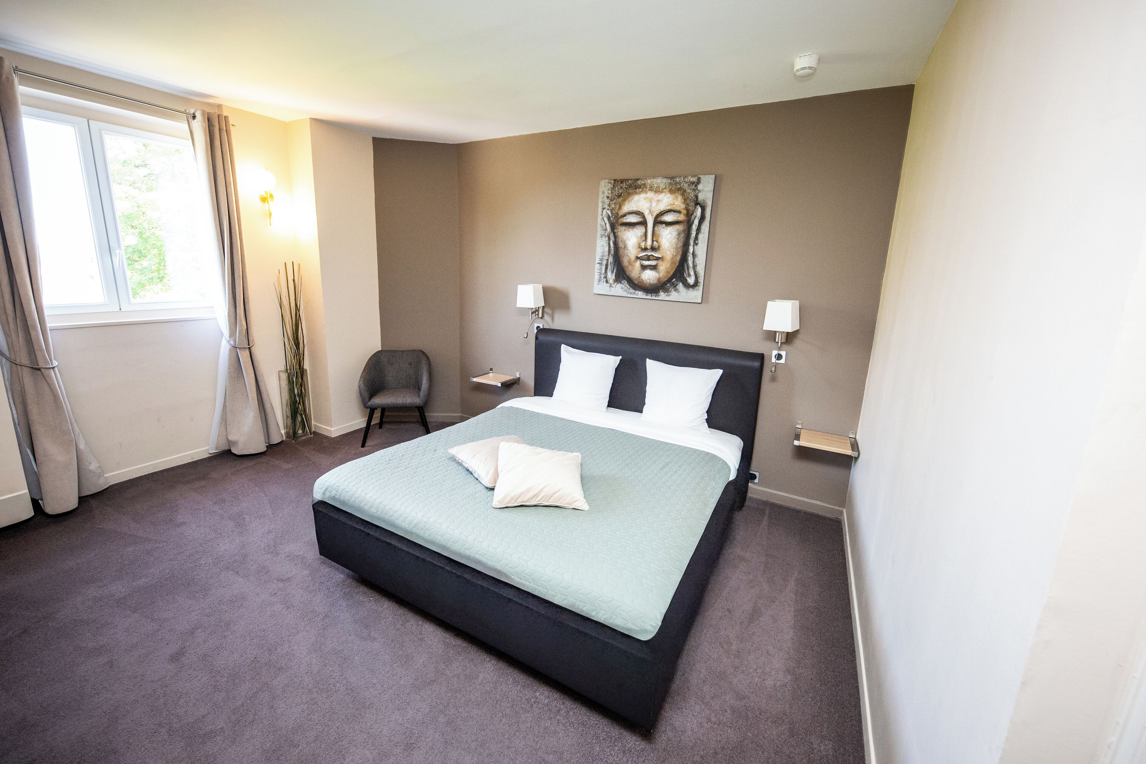 Een slaapkamer met een groot bed, twee bedlampen, een Boeddha-schilderij aan de muur en een raam met gordijnen.