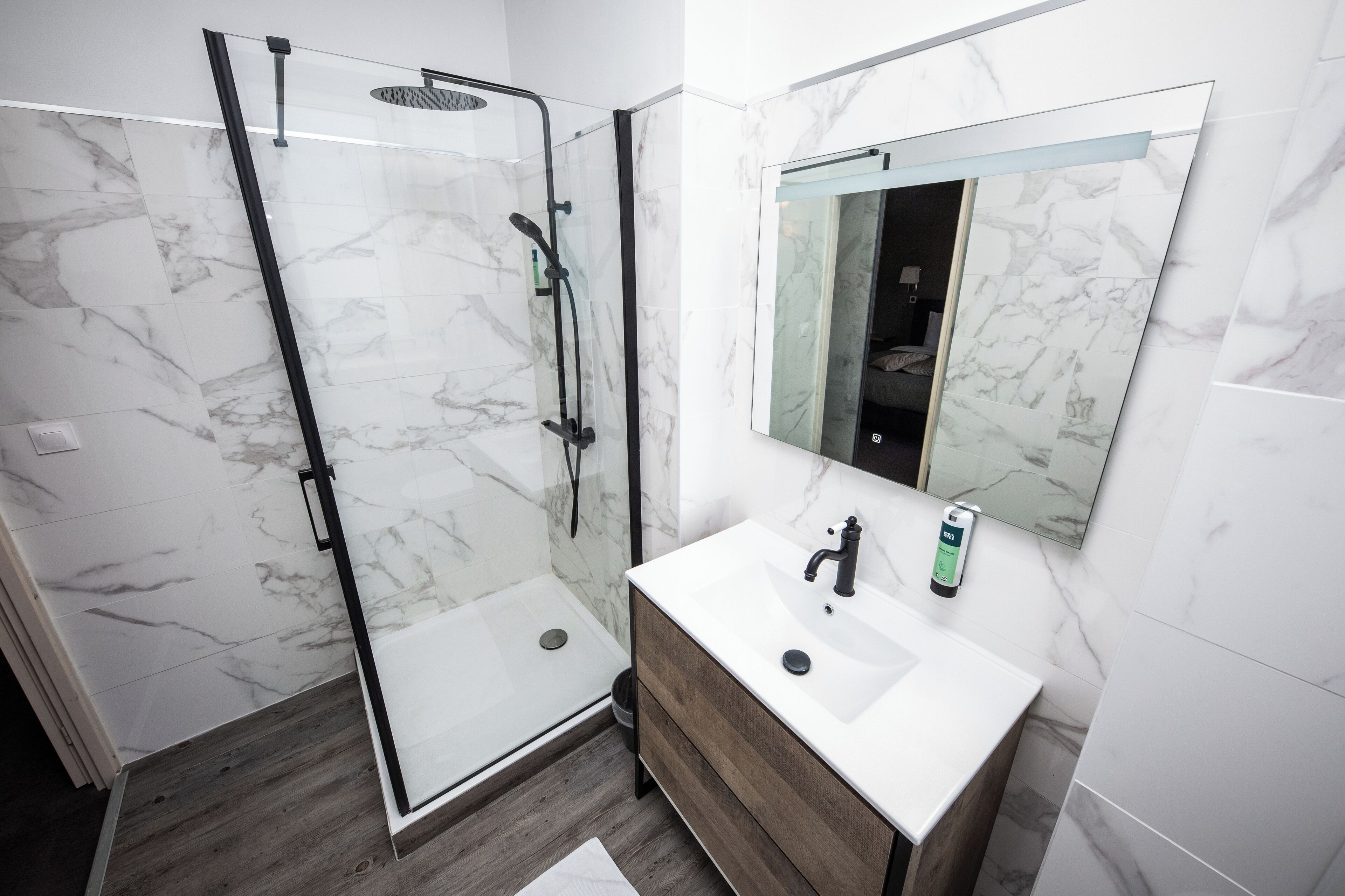 Un baño moderno con ducha envuelta en vidrio, lavamanos blanco sobre tocador de madera y pared con azulejos de mármol.