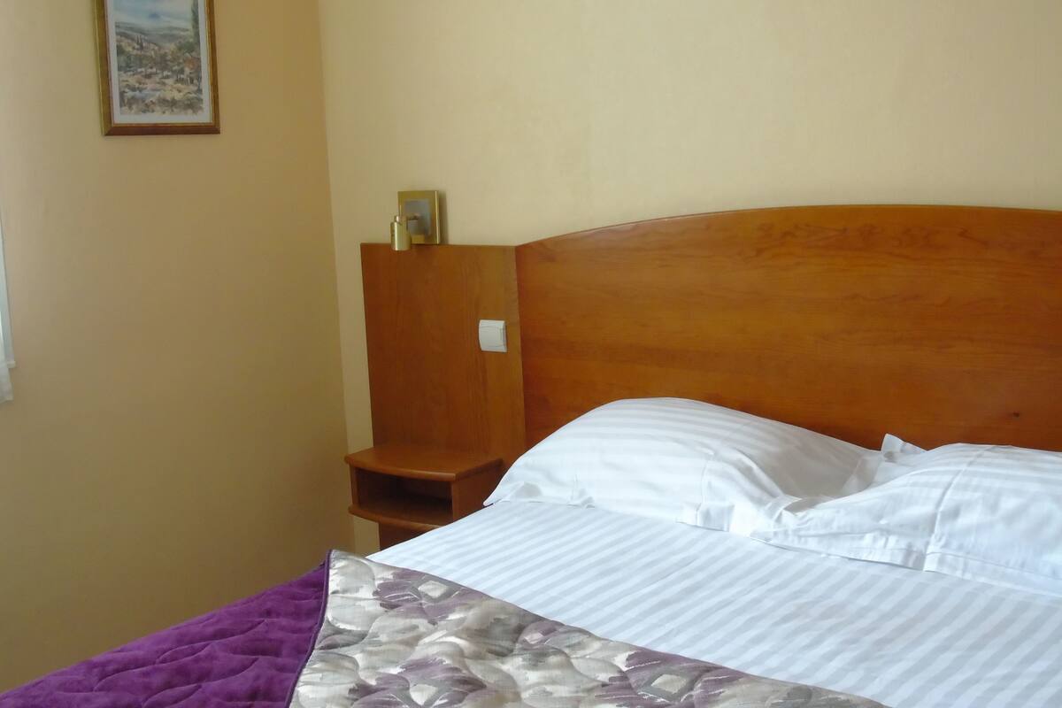 A hotel room with a wooden headboard, a bed with white linens, a purple blanket, and a small wooden nightstand with a lamp.
