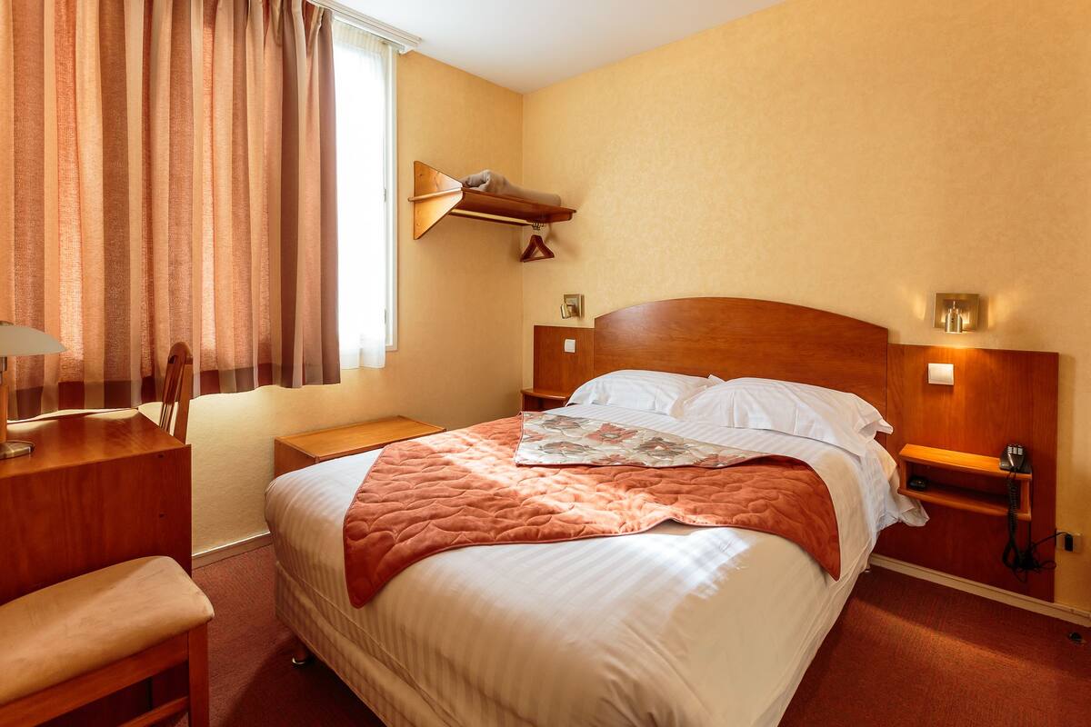 A hotel room with a large bed, a bedside table, a lamp, a chair, and a window with curtains.