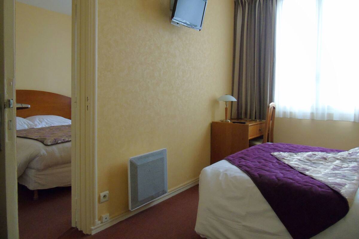 A hotel room with a bed, a television mounted on the wall, a desk with a lamp, and a window with curtains.