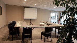A conference room with a long table, chairs, a projector screen, and a plant in the foreground. - Hotel De la Place (Les Monts d'Aunay)