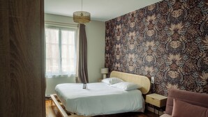 A bedroom with a bed, a bedside table, a lamp, a chair, and a patterned wallpaper. - Hotel De la Place (Les Monts d'Aunay)