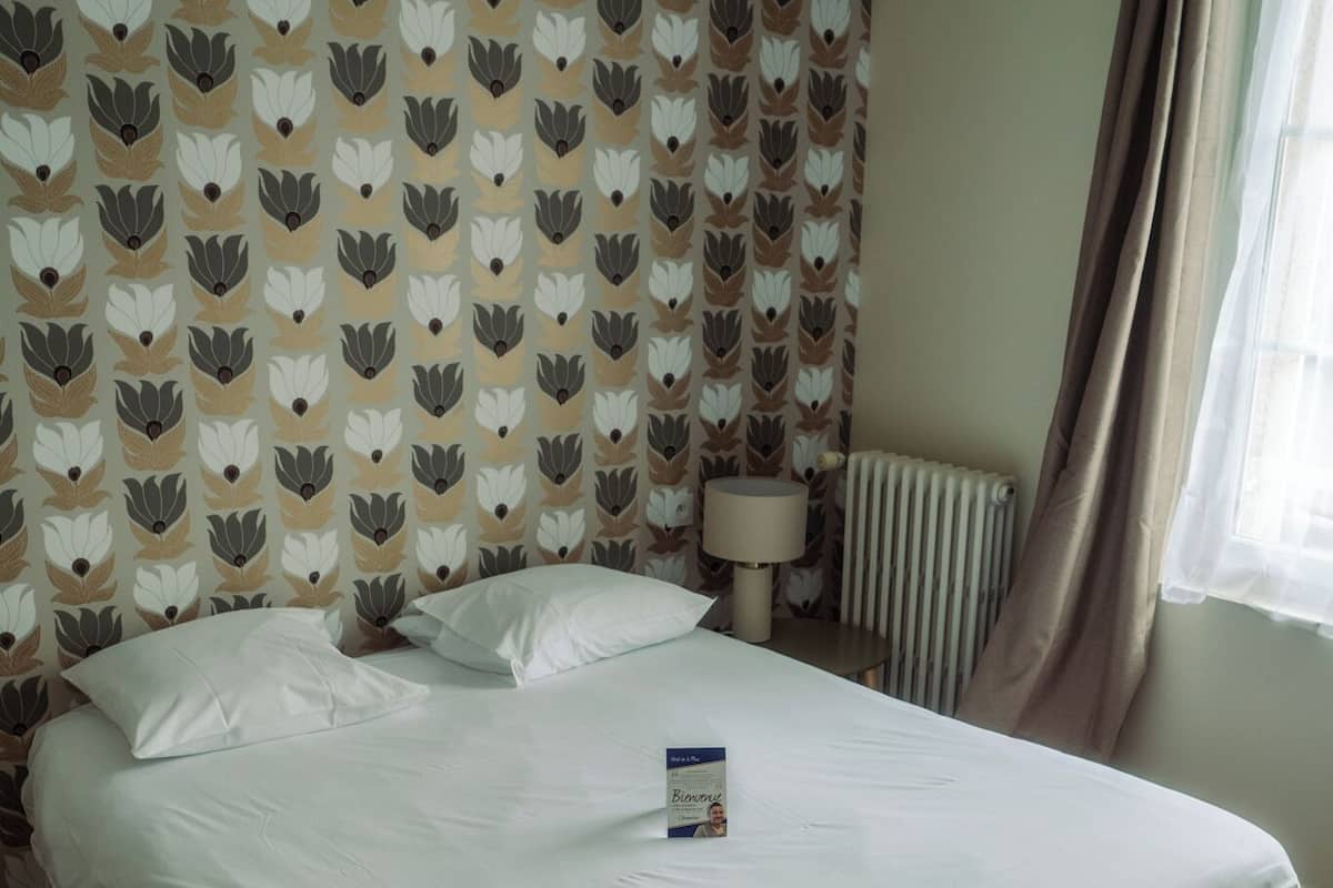 Ein ordentlich bezogenes Bett in einem Zimmer mit strukturiertem Wandpapier, einer Nachttischlampe und einem Fenster mit Vorhängen.