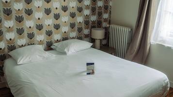 A neatly made bed in a room with patterned wallpaper, a bedside lamp, and a window with curtains.