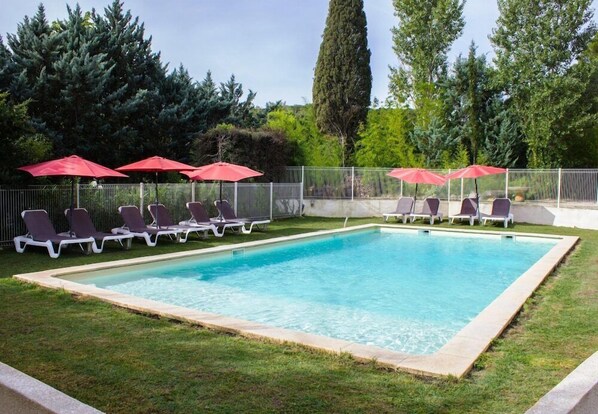 A rectangular swimming pool with a tiled edge, surrounded by a grassy area. There are multiple lounge chairs and red umbrellas arranged around the pool. - Le Colombier (Remoulins)
