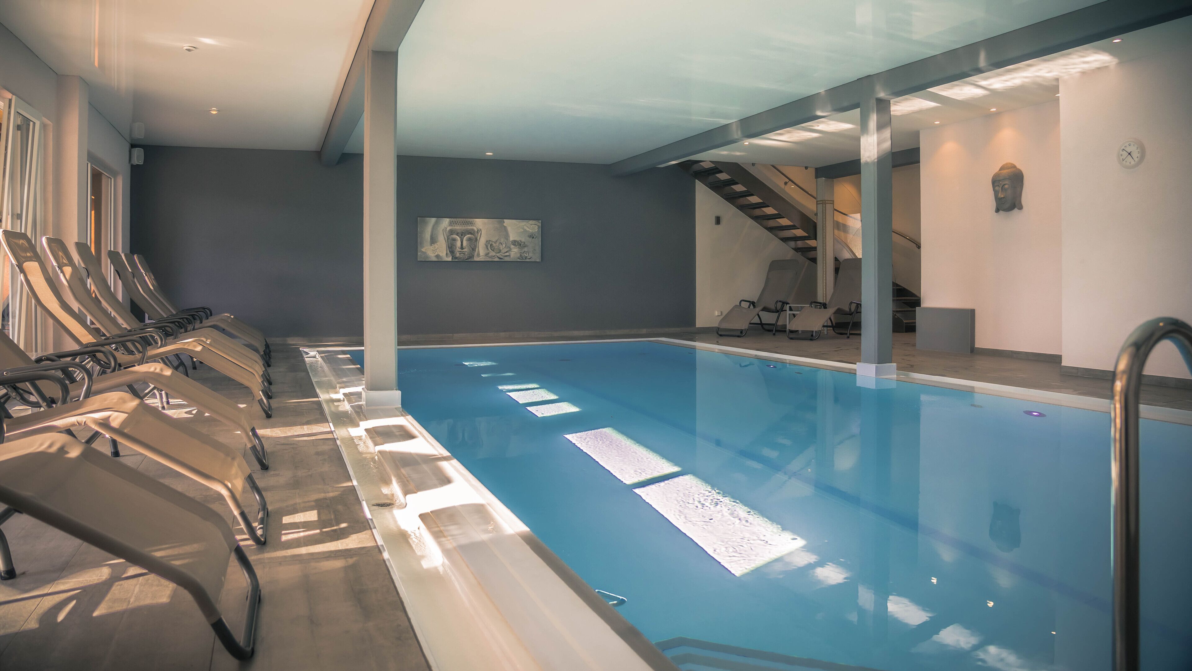 A swimming pool with sunlit water, lounge chairs, and a Buddha statue.