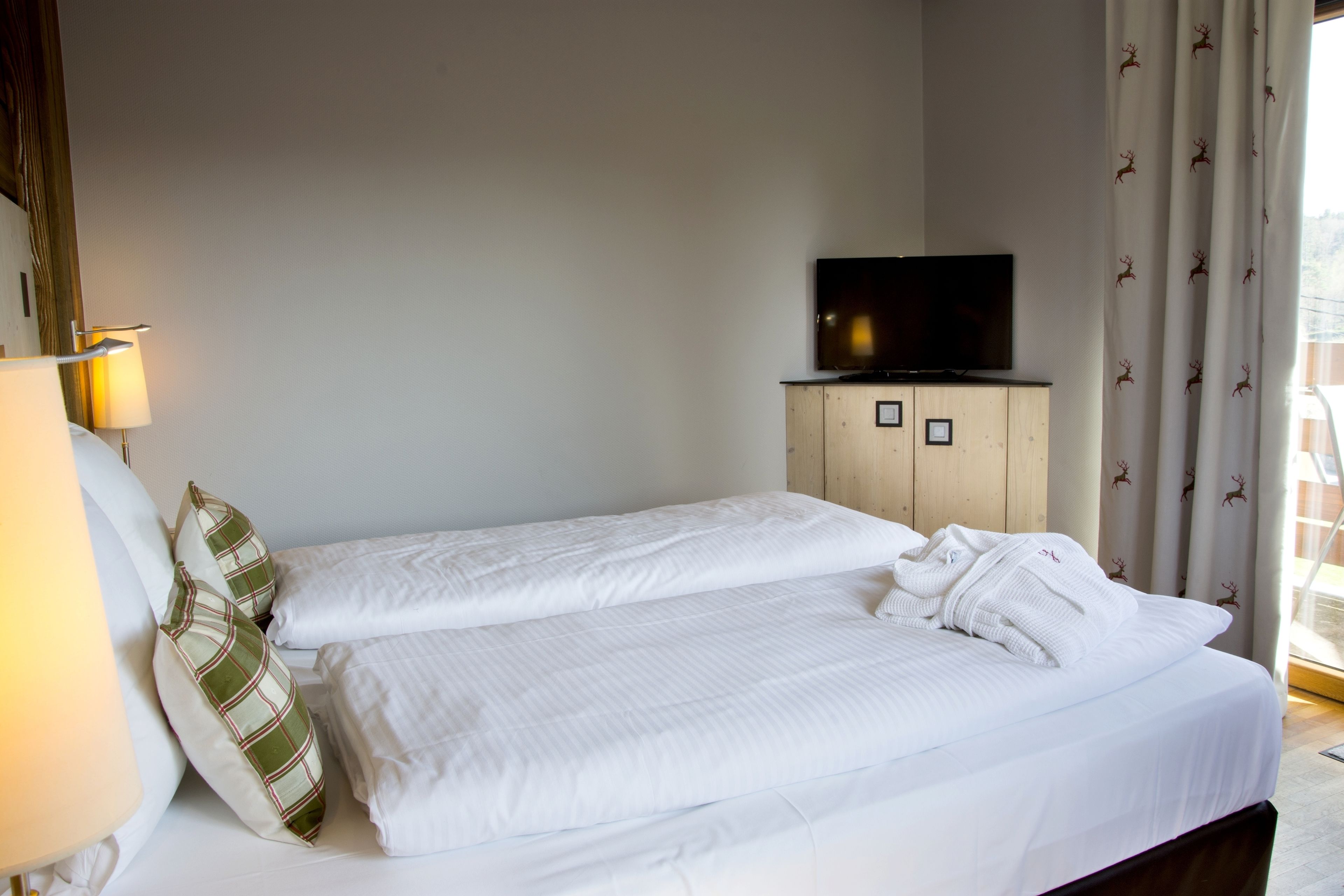 Habitación de hotel con dos camas, un televisor sobre un mueble de madera y una ventana con cortinas.