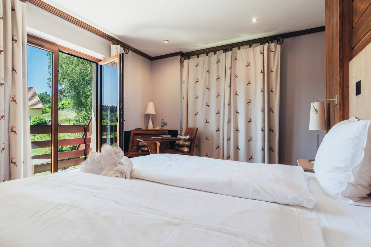 A hotel room with a large bed, a desk with a chair, a lamp, and a view of greenery outside.