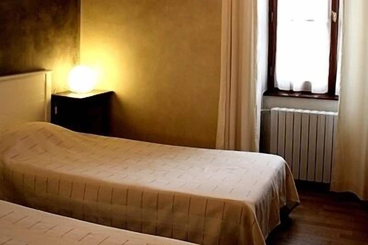 A room with two beds, a bedside table with a lamp, a radiator, and a window with curtains.