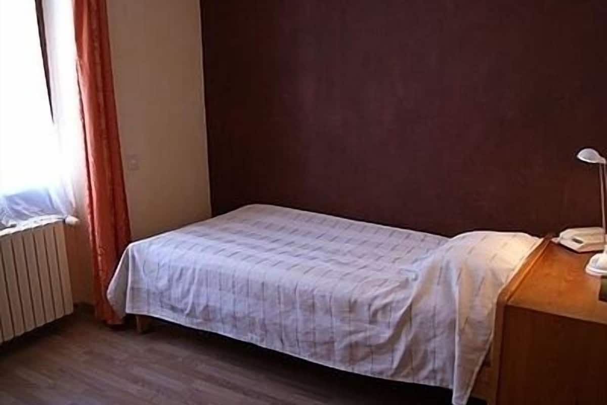 A small, single-bed room with a wooden floor, a bedside table with a lamp, and a window with curtains.